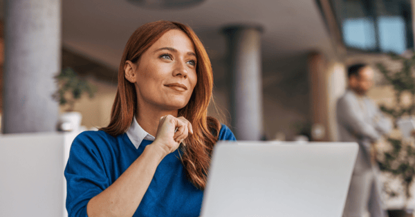 Red haired woman in a blue jumper looking off into the distance, sat at a desk with her laptop.