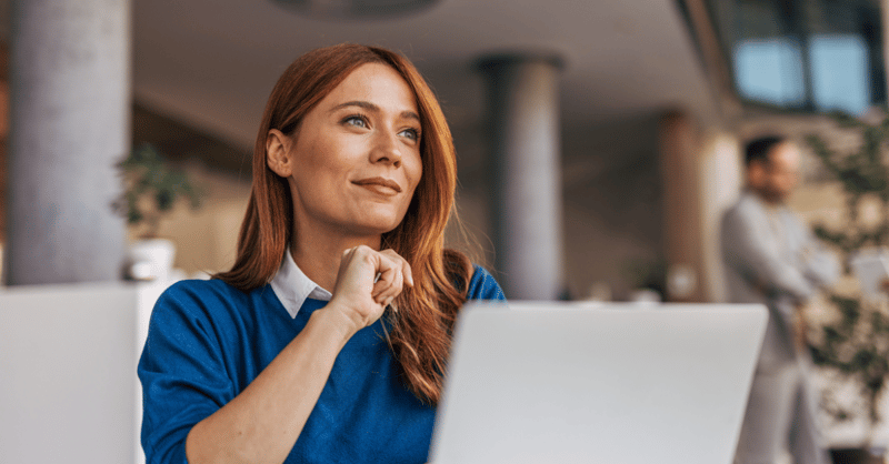 Red haired woman in a blue jumper looking off into the distance, sat at a desk with her laptop.