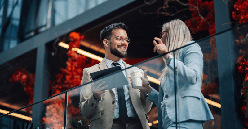 Two business colleagues are standing on a balcony, having a conversation about work while holding a tablet and a coffee cup