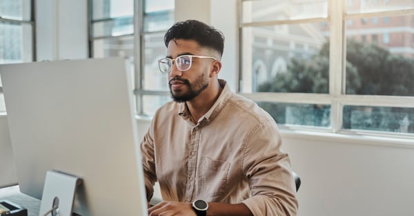 Young male employee wearing glasses and beige shirt sat at computer