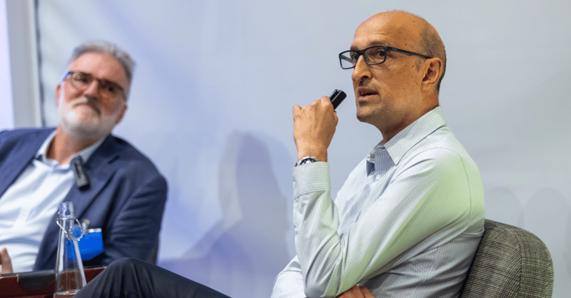 Matthew Syed, holding a microphone and speaking to the audience, with Jacques Quinio sat beside him