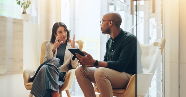 Male and female employee in conversation, sat in a small meeting room. The man is holding a tablet. 