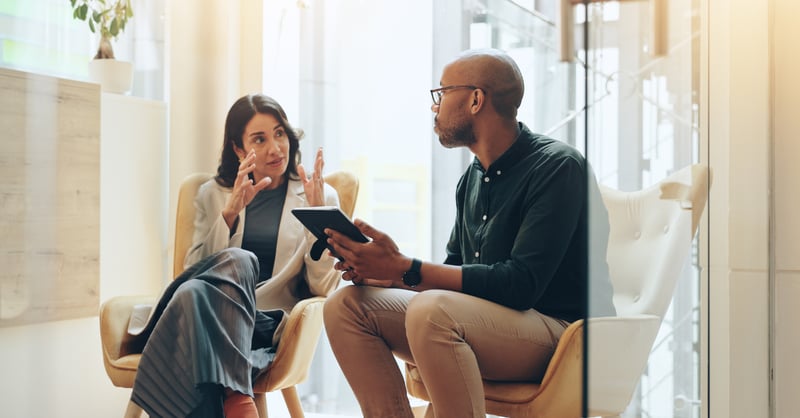Male and female employee in conversation, sat in a small meeting room. The man is holding a tablet. 