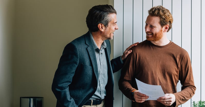 Two men in conversation and laughing. One is wearing a blue blazer and shirt, the other an orange jumper.