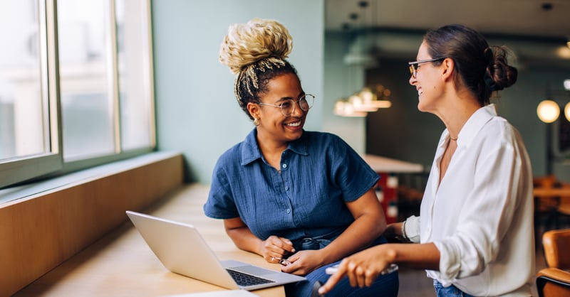Two women are seated in a bright indoor setting, engaging in conversation near a laptop