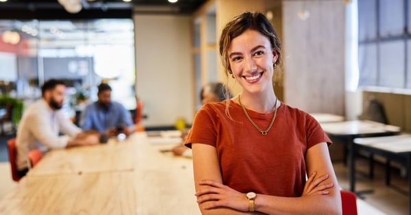 Woman in orange top smiling at camera while colleagues work at a desk behind her