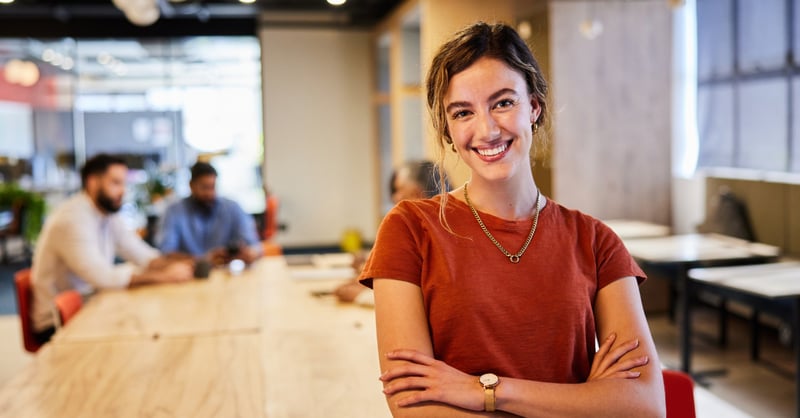 Woman in orange top smiling at camera while colleagues work at a desk behind her