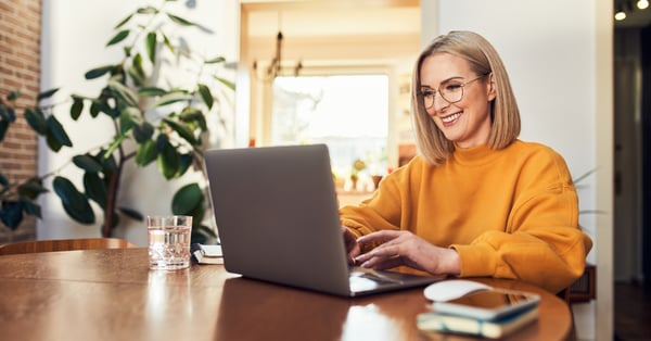 Woman in yellow jumper sat at a table and smiling at her laptop